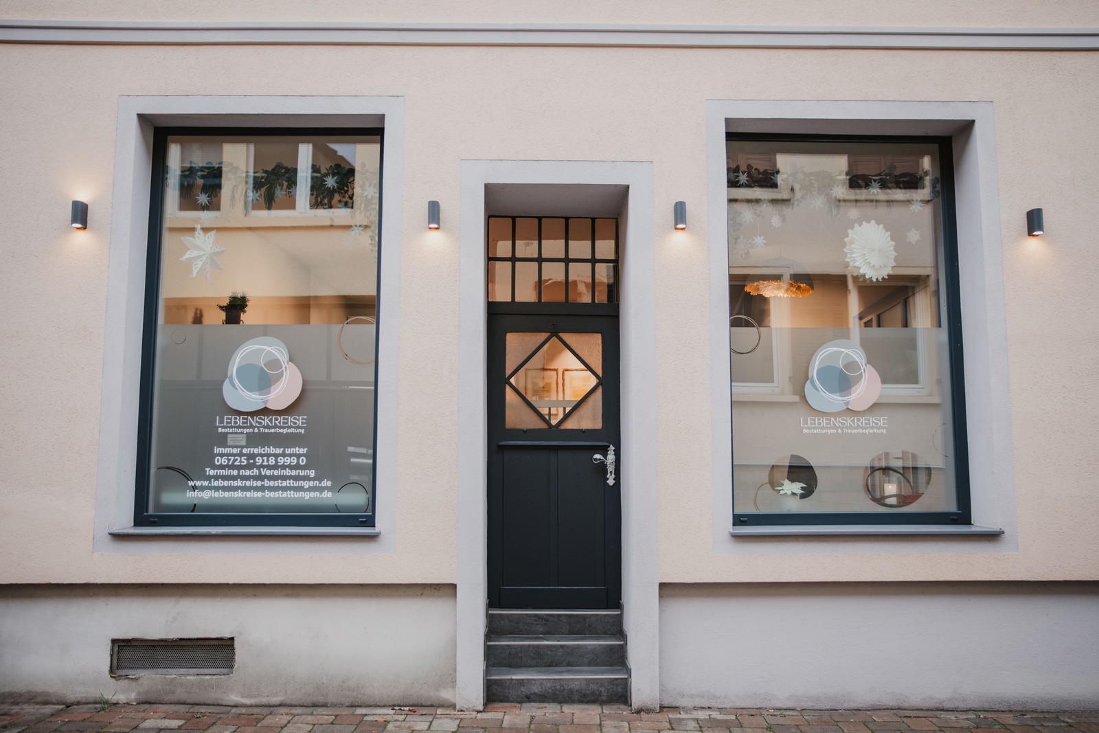 Unsere Filiale von Lebenskreise Bestattungen in Gau-Algesheim mit schwarzer Tür, Schaufenstern und Deko.