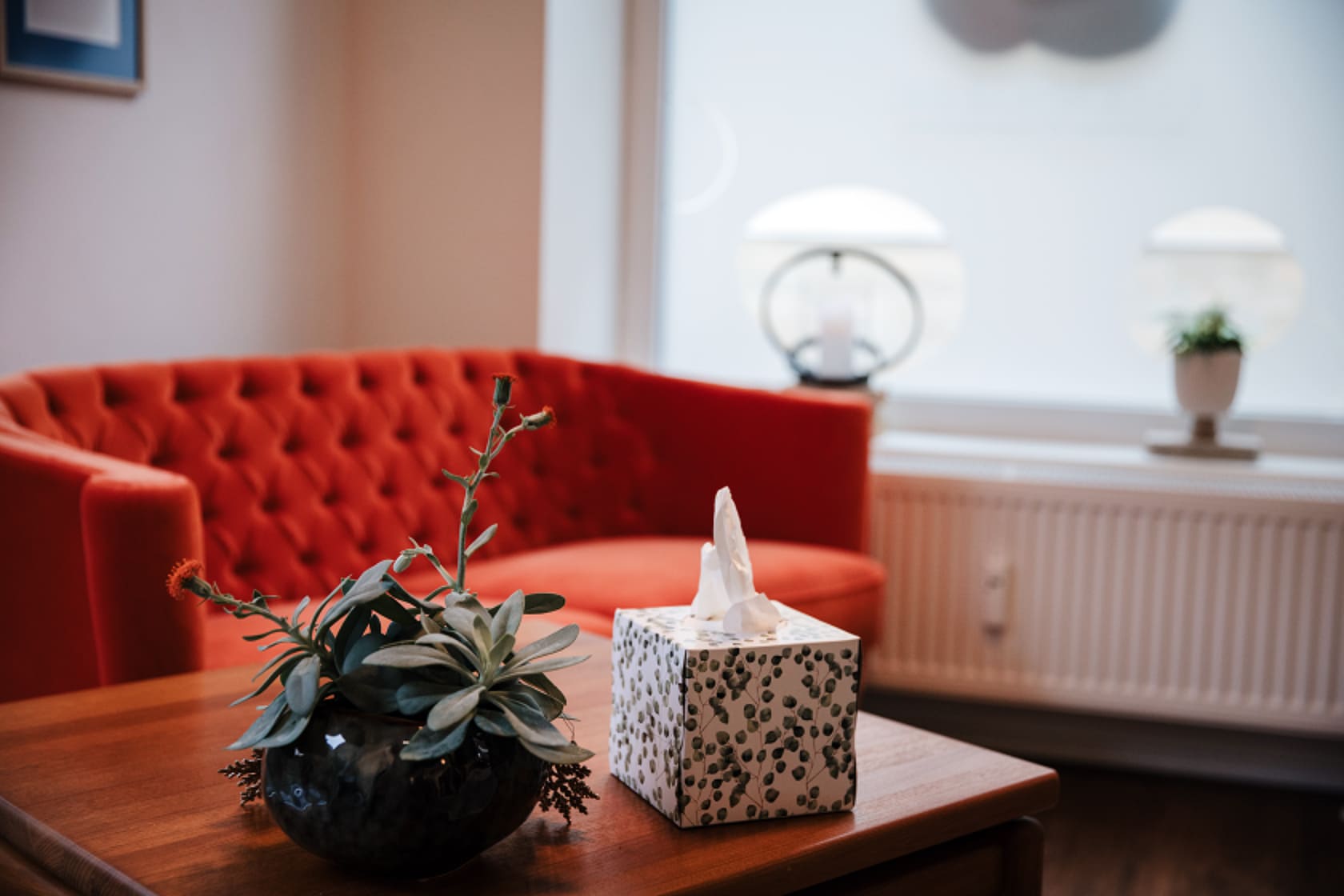 Gemütlicher Besprechungsraum bei Lebenskreise Bestattungen: Rotes Sofa am Holztisch mit Pflanze, Taschentüchern und warmem Licht.