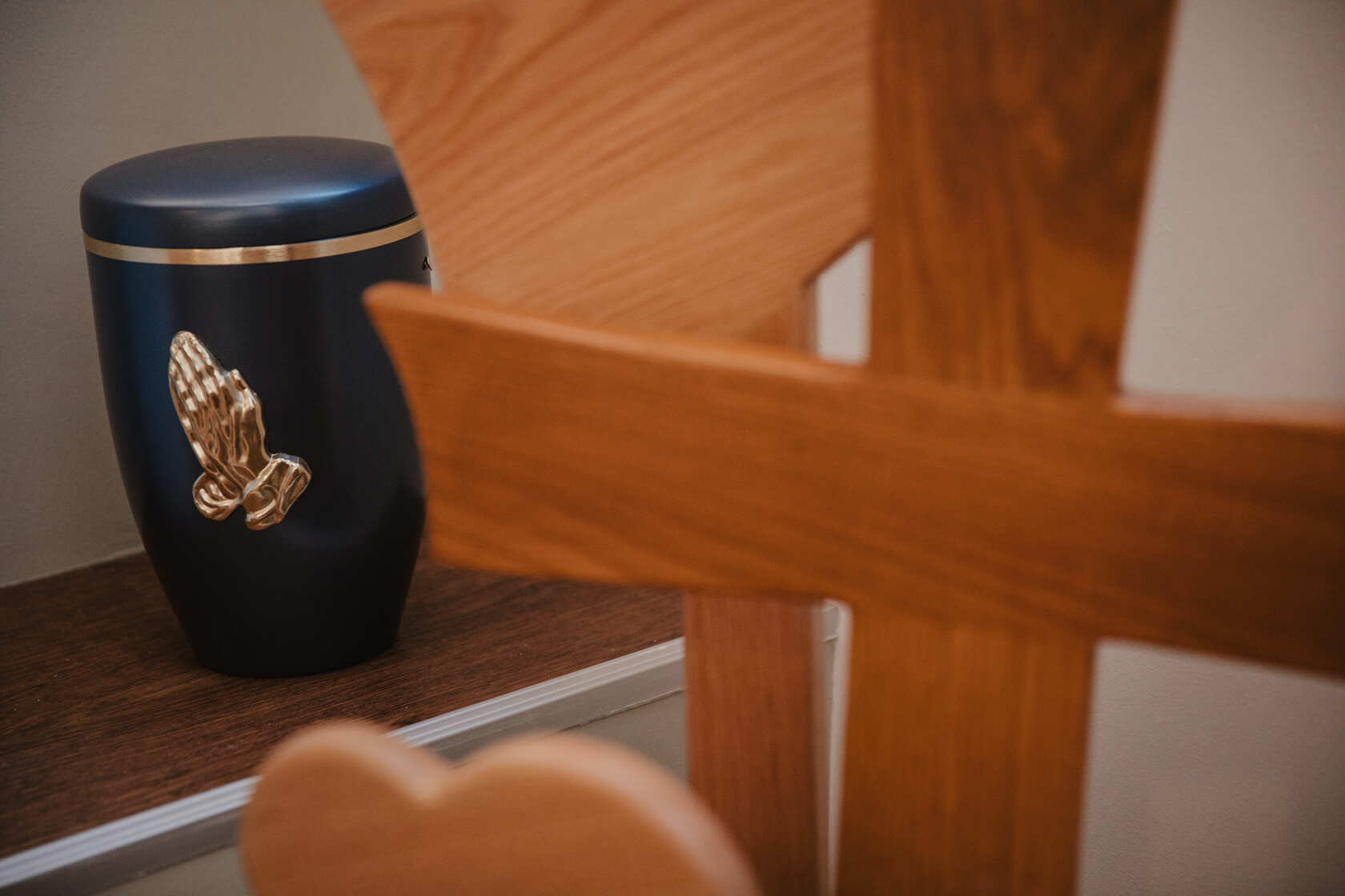 Schwarze Urne mit goldenen betenden Händen, neben einem Holzkreuz - bei Lebenskreise Bestattungen in Gau-Algesheim.