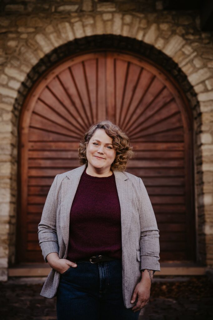 Katharina Kluge, mit lockigen Haaren im grauen Blazer und bordeauxroten Pullover, vor einer Holztür aus Steinmauer stehend.